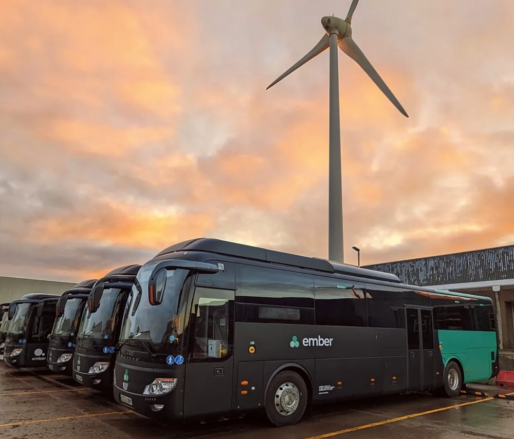 Row of Ember electric coaches parked up in front of a wind turbine