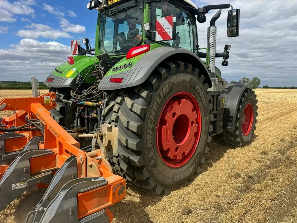 Image of a green tractor ploughing a field with red wheels. It is viewed from a rear angle.